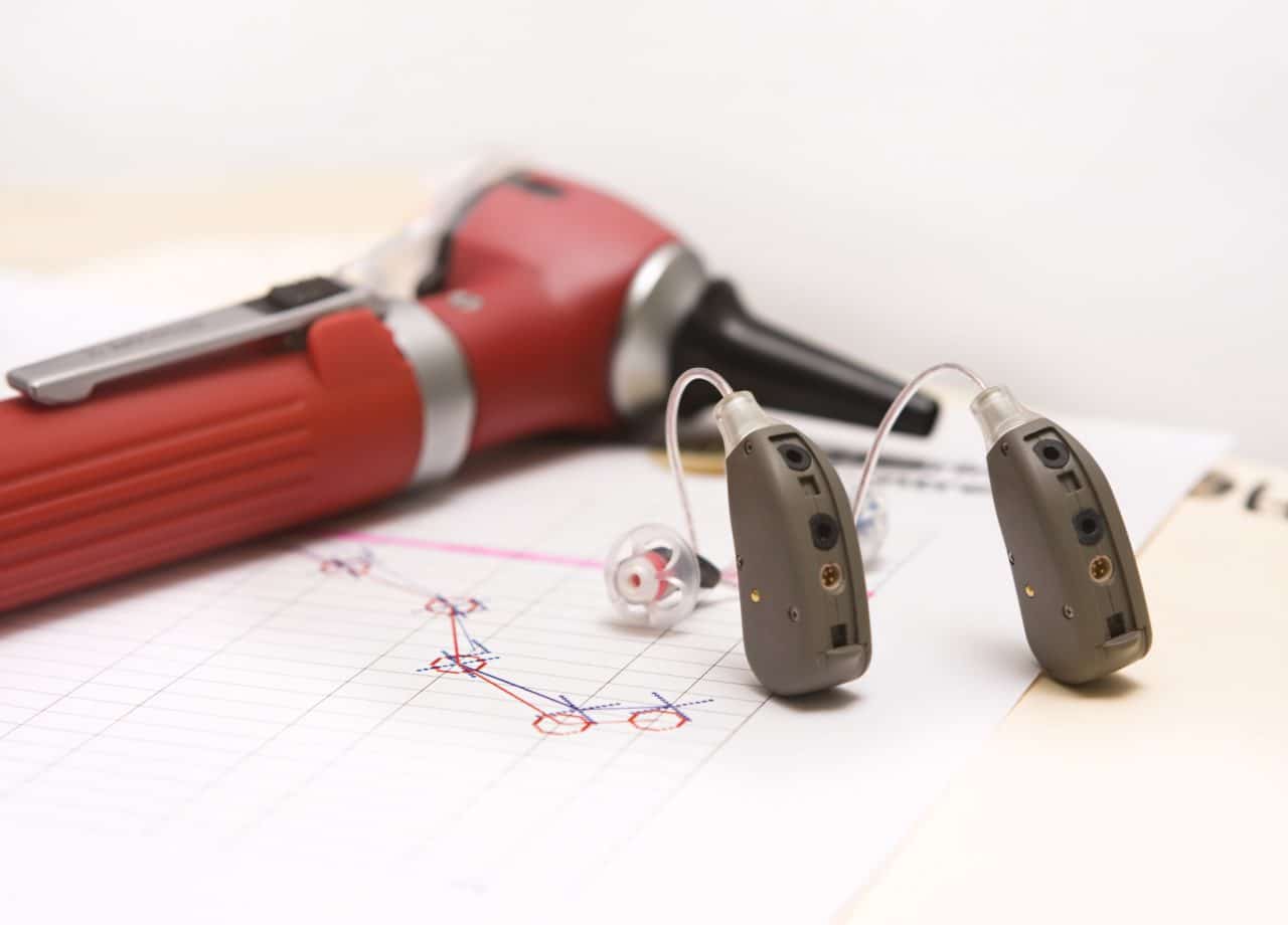 A pair of modern hearing aids on a medical chart showing a hearing loss of high frequencies. An otoscope lies in the background.